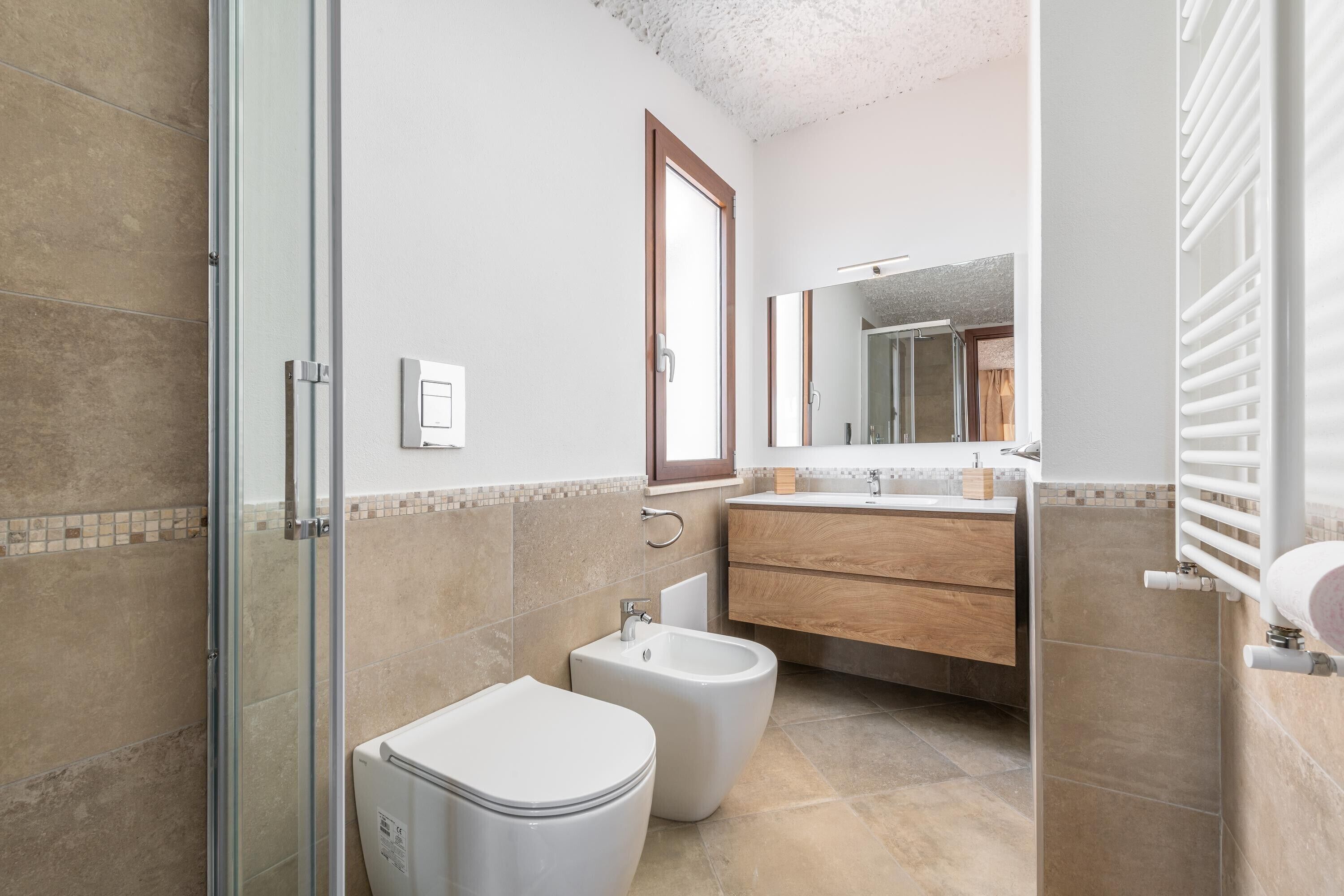 A bathroom with a glass shower enclosure, a bidet, a toilet, a sink with a mirror, and a towel rack.