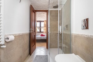 A bathroom with a shower, toilet, and a visible bed in the adjacent room.