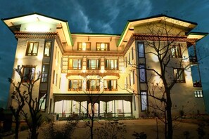 Um edifício grande e com vários andares, fachada simétrica, diversas janelas e entrada central coberta.