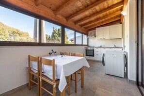 Un comedor con mesa y sillas, una cocina con armarios y electrodomésticos, y ventanas amplias con vistas a árboles.