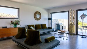 Un salon moderne avec un canapé gris, des coussins verts, un miroir rond et une vue sur l’extérieur grâce à de grandes fenêtres.