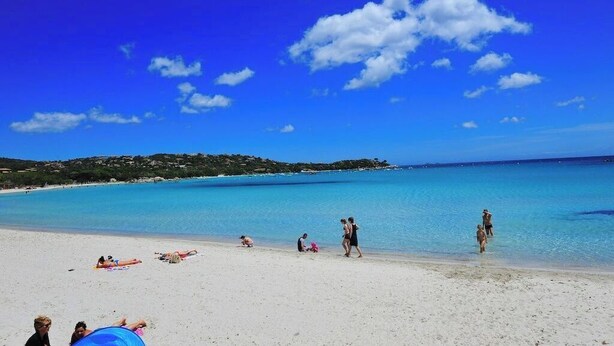 Une plage avec une eau d’un bleu limpide, des gens qui prennent des bains de soleil et une côte visible au loin.