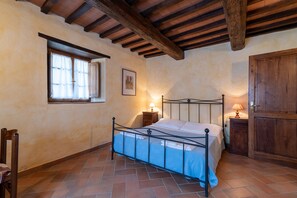 Un dormitorio con estructura de cama de metal, vigas de madera en el techo, una ventana con cortinas, una mesita de noche con lámpara y un armario de madera.