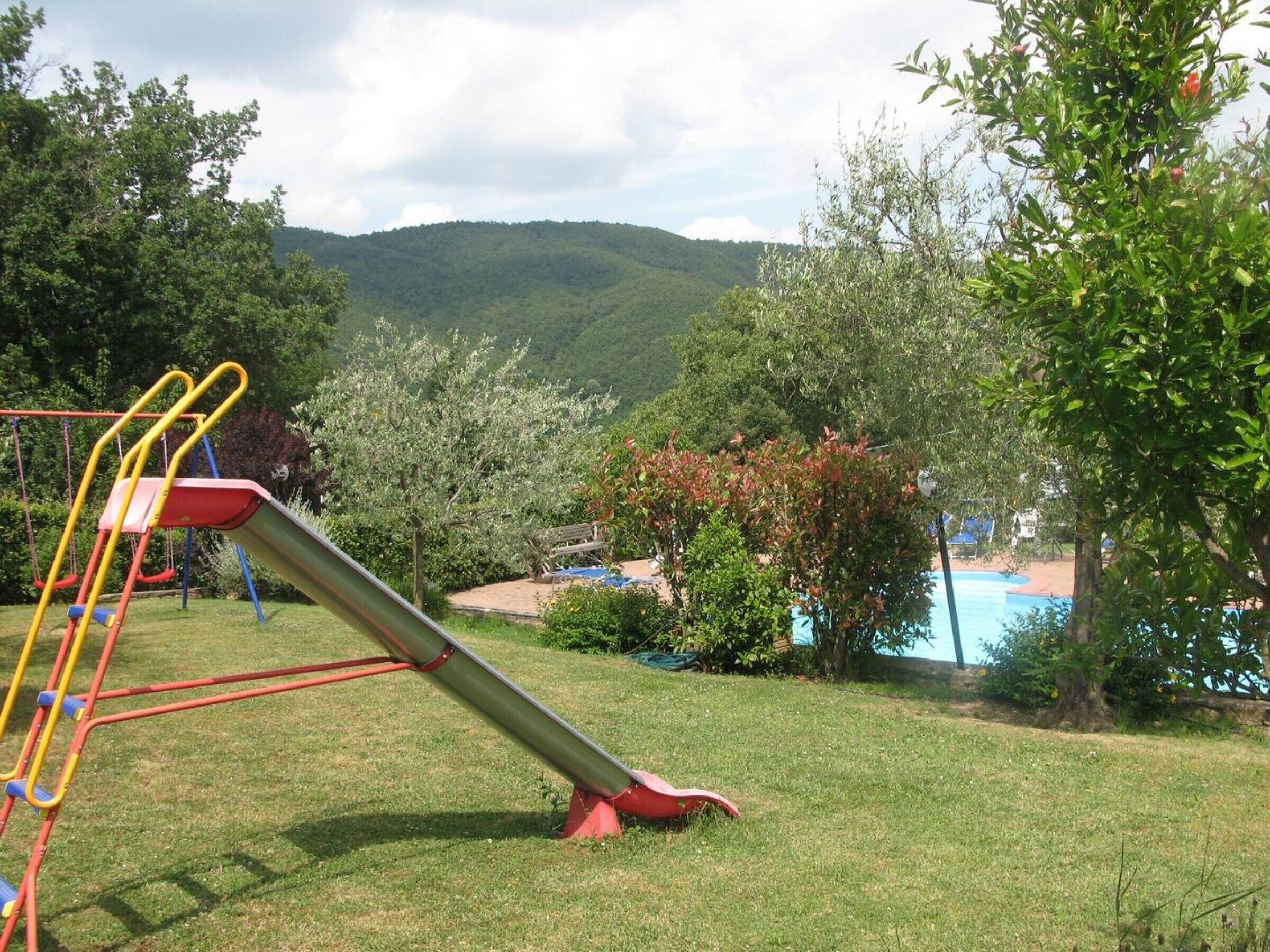 Ein Spielplatz mit Rutsche und Schaukeln, umgeben von viel Grün und einem Schwimmbecken.