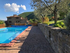 Ein Swimmingpool mit Steinmauer und Holzzaun, ein Steinhaus mit Ziegeldach und eine bergige Landschaft im Hintergrund.