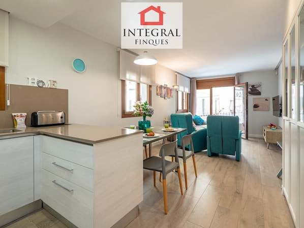 A modern kitchen with a toaster, a clock, and a welcome bag. There is a dining area with teal chairs and a table.