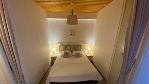 Un couloir étroit mène à un lit doté d’une tête de lit en bois, de deux tables de chevet avec des lampes et d’un luminaire fixé au plafond.
