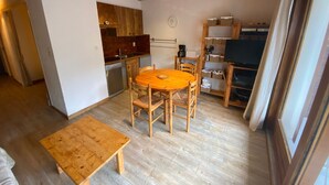 Un espace de vie compact comprenant une table à manger en bois, des chaises, un petit coin cuisine et un téléviseur posé sur une étagère.