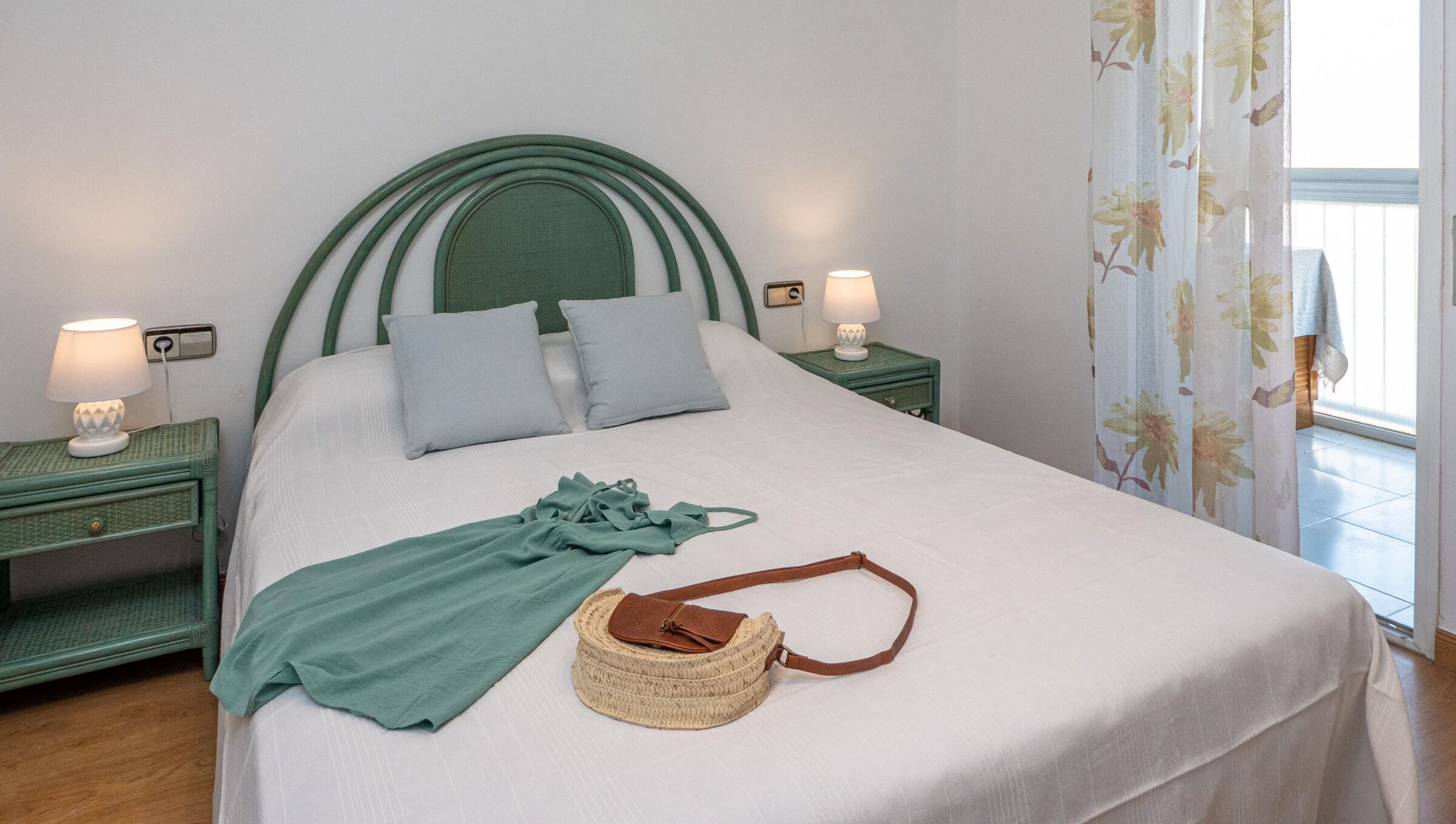 Un lit bien fait, avec une tête de lit verte, deux tables de chevet avec des lampes, un sac en tissu tressé et un tissu vert sur le lit.