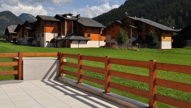 A row of wooden chalets with sloped roofs, set against a backdrop of green fields and mountains.