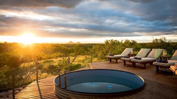 Et poolområde med liggestole og jacuzzi, omgivet af en træterrasse med udsigt over et smukt landskab.