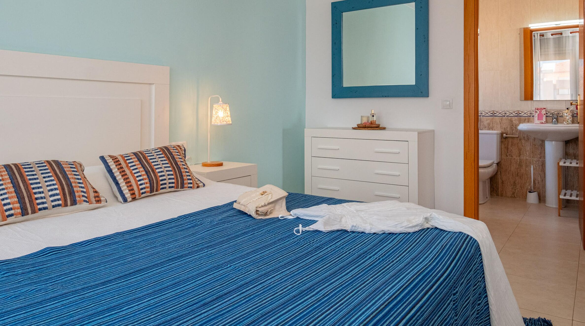 A hotel room with a bed, a nightstand, a dresser, and a bathroom with a sink and mirror.