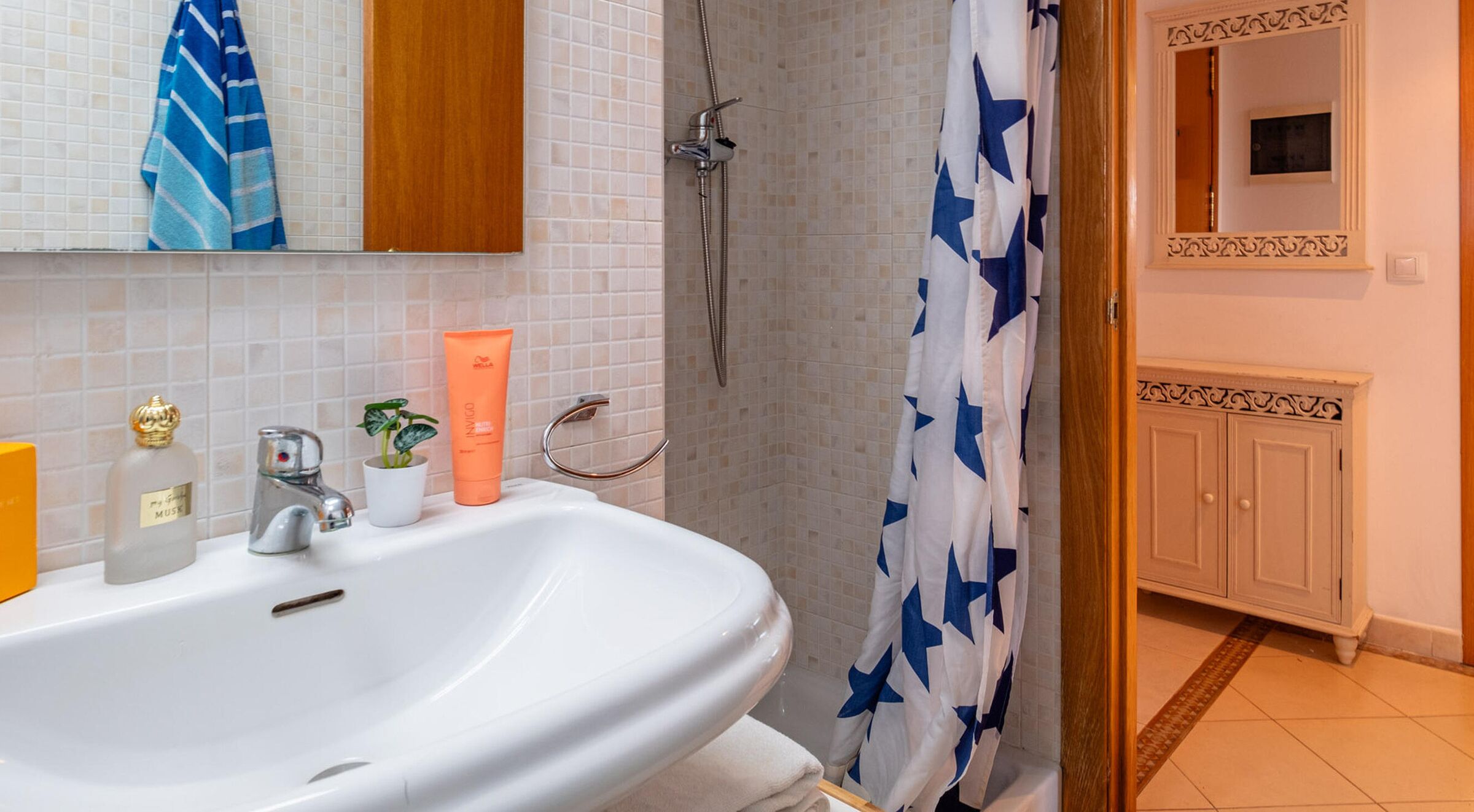 Une salle de bain avec un lavabo blanc, une douche avec un rideau de douche bleu et blanc, un miroir et une petite plante en pot.