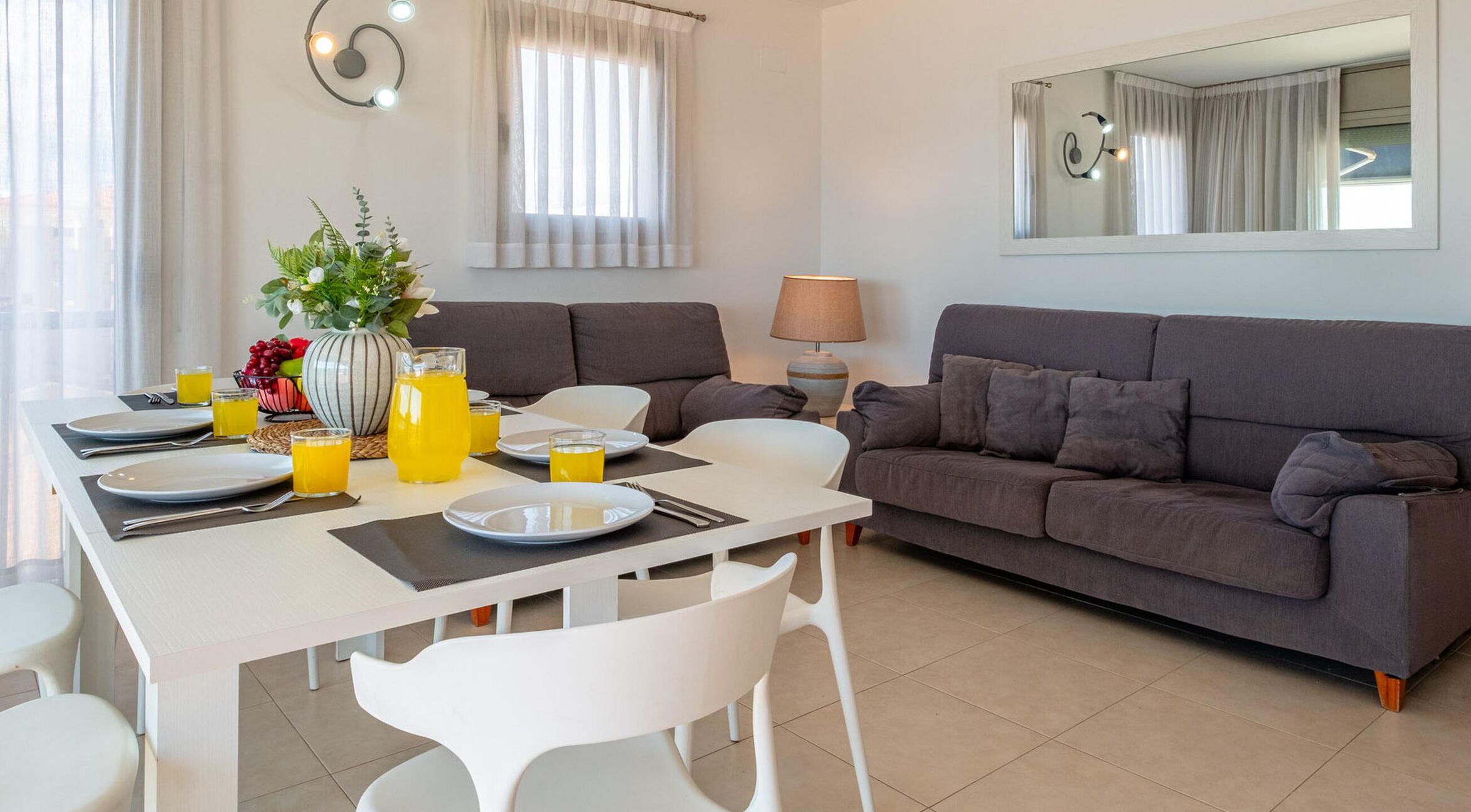 Een moderne woonkamer met een eetgedeelte, een grijze bank, witte stoelen, een tafel gedekt voor ontbijt, en een grote spiegel aan de muur.