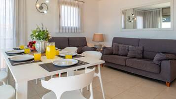 Een moderne woonkamer met een eetgedeelte, een grijze bank, witte stoelen, een tafel gedekt voor ontbijt, en een grote spiegel aan de muur.