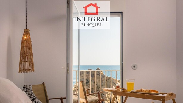 Une chambre avec un lit, une table de chevet sur laquelle se trouve un verre de jus d’orange, une lampe en osier et une vue sur la mer, visible par la porte d’un balcon.