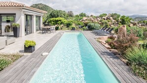 Et rektangulært pool med træterrasser på hver side, omgivet af grønt og et bjerglandskab.