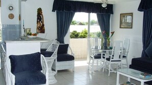 A spacious living area with white wicker furniture, a glass-top table, and a balcony with blue curtains. - Luxurious beach front self-catering apartment in the heart of Ocho Rios, Jamaica (Ocho Rios)