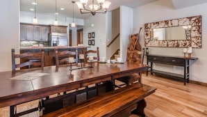 Un grand espace à manger avec une longue table en bois et des bancs, une cuisine à l’arrière-plan et un escalier menant à un autre niveau.