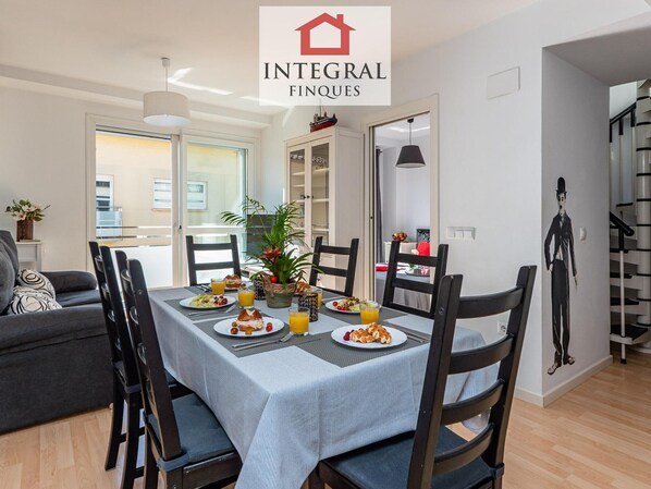 A dining area with a table set for four, surrounded by chairs. There is a plant on the table, and the room has a modern design with a staircase in the background.