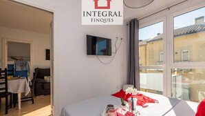 A hotel room with a bed, a dining area with a table and chairs, a TV mounted on the wall, and a balcony with a view of buildings. - Stunning Duplex 200m from Platja Gran, 3BR, Balcony, Terrace, Parking, WiFi, A/C, Lift (Palamós)