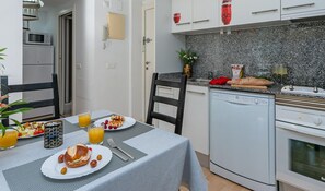 Una cocina moderna con armarios blancos, encimera de granito y horno empotrado. Hay una mesa de comedor con alimentos para el desayuno y una silla.