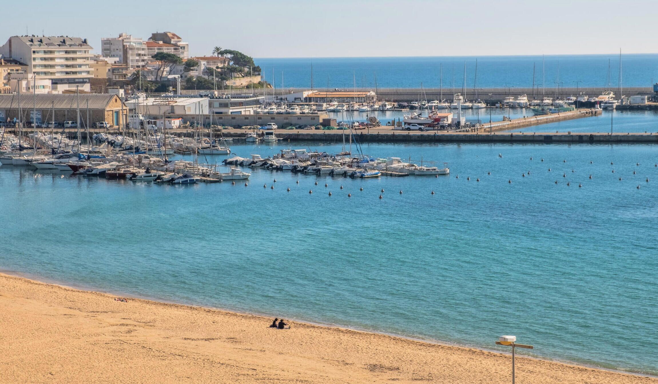 Un port de plaisance sur la côte, avec des bateaux amarrés, une plage de sable et des bâtiments en arrière-plan.