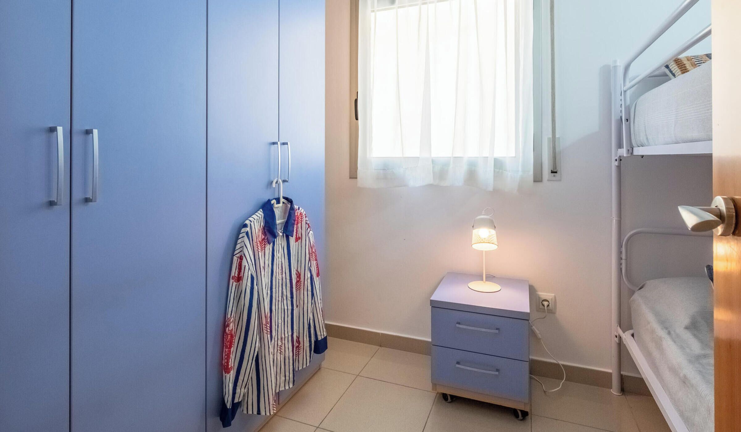 Een slaapkamer met een stapelbed, een kledingkast, een nachtkastje met een lamp en een hangende kledingrek.