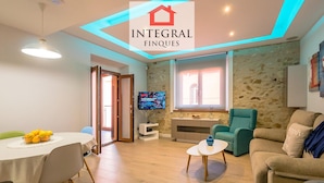A modern living room with a stone wall, a television, a sofa, a coffee table, and a dining area with a table and chairs. - Modern Apartment, 2 Bedrooms, 2 Bathrooms, Balcony, Parking, 100m from Beach (Palamós)