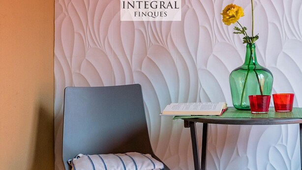 A chair with a cushion, a table with a book and a vase with flowers, and a wall with a logo. - Modern Apartment, 2 Bedrooms, 2 Bathrooms, Balcony, Parking, 100m from Beach (Palamós)