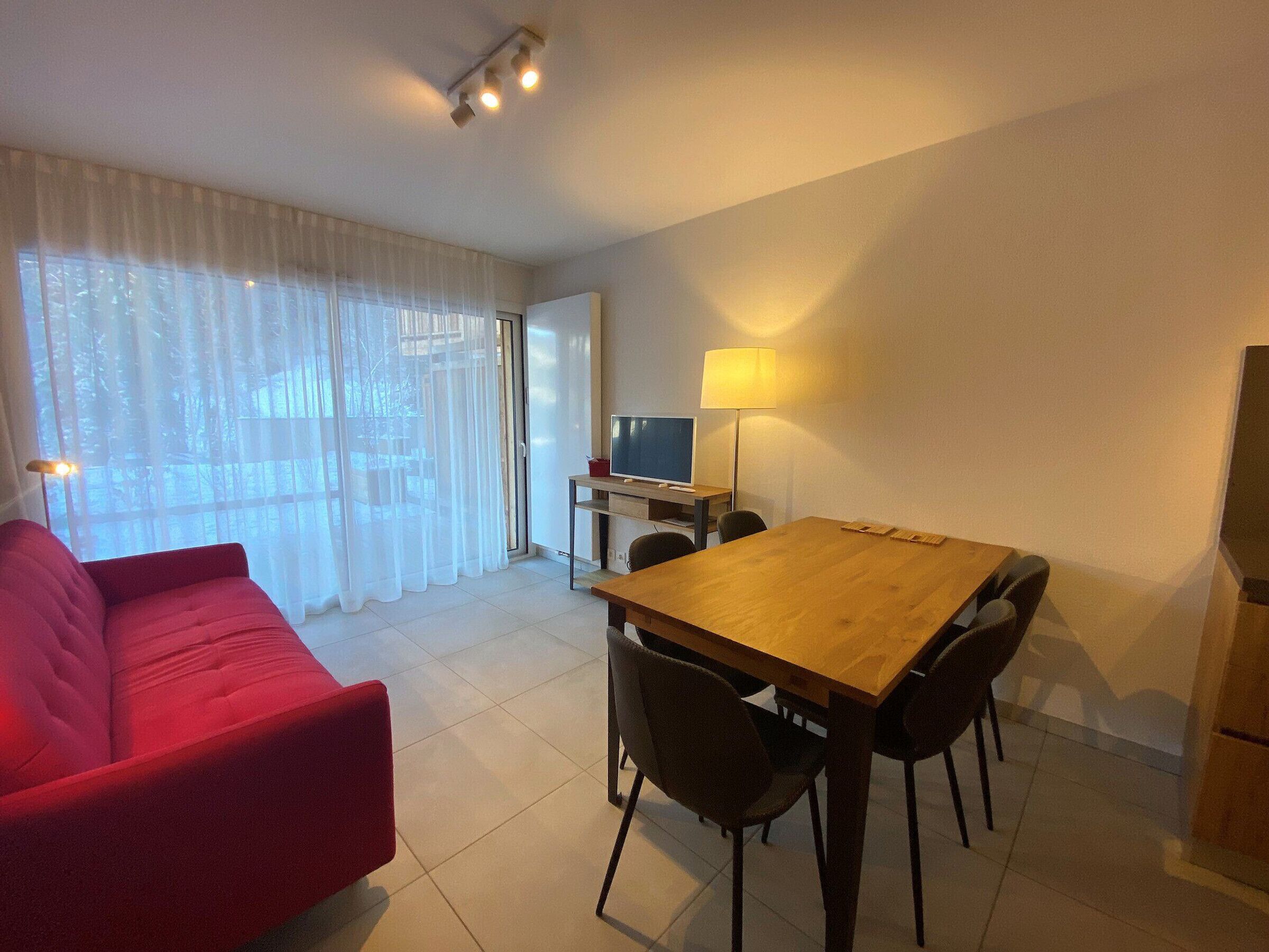 Uma sala de estar moderna com um sofá vermelho, uma mesa de jantar de madeira com quatro cadeiras, uma televisão e um abajur.