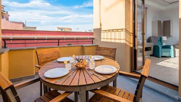 Una mesa redonda de madera con platos, cubiertos y un bol de fruta en un balcón con vista a la ciudad.