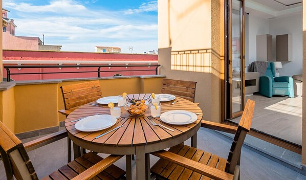 A round wooden table with plates, cutlery, and a bowl of fruit on a balcony with a city view.