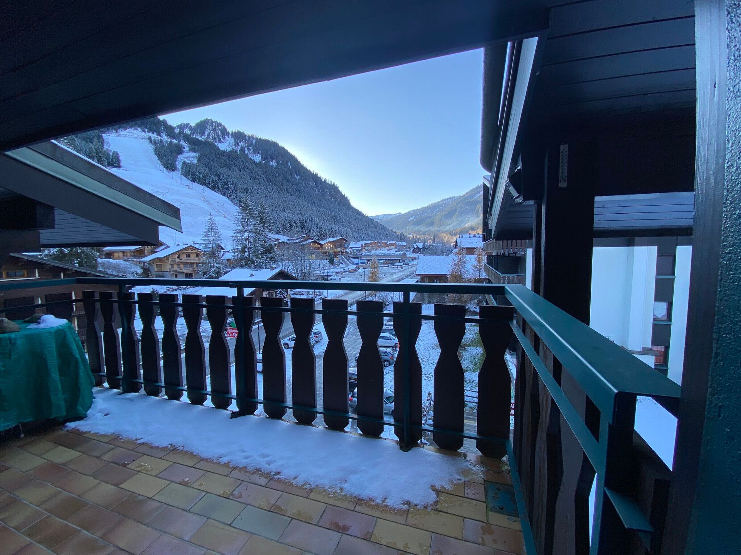 A balcony with a view of a snowy mountain landscape, a tiled floor, and a green tablecloth.