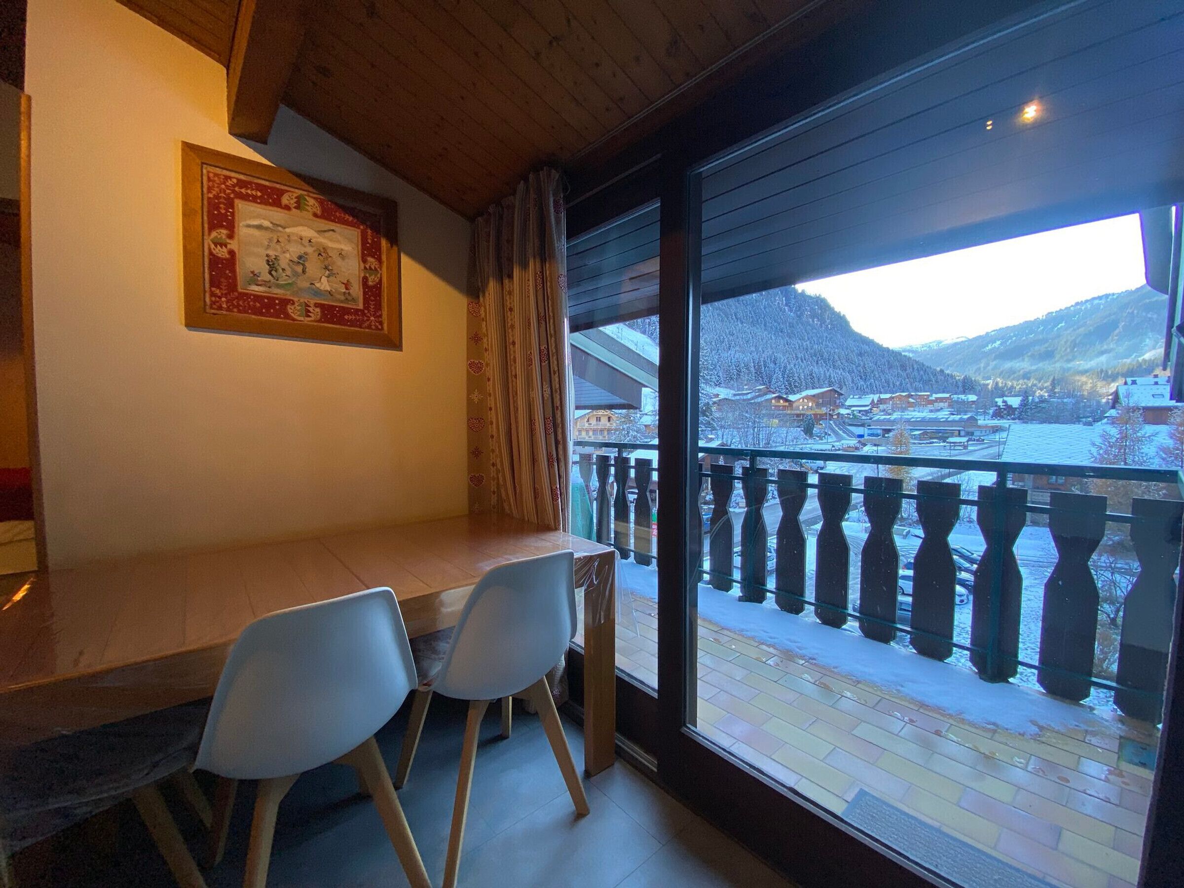 A dining area with a table and chairs, a framed picture on the wall, and a view of a snowy landscape through a balcony.