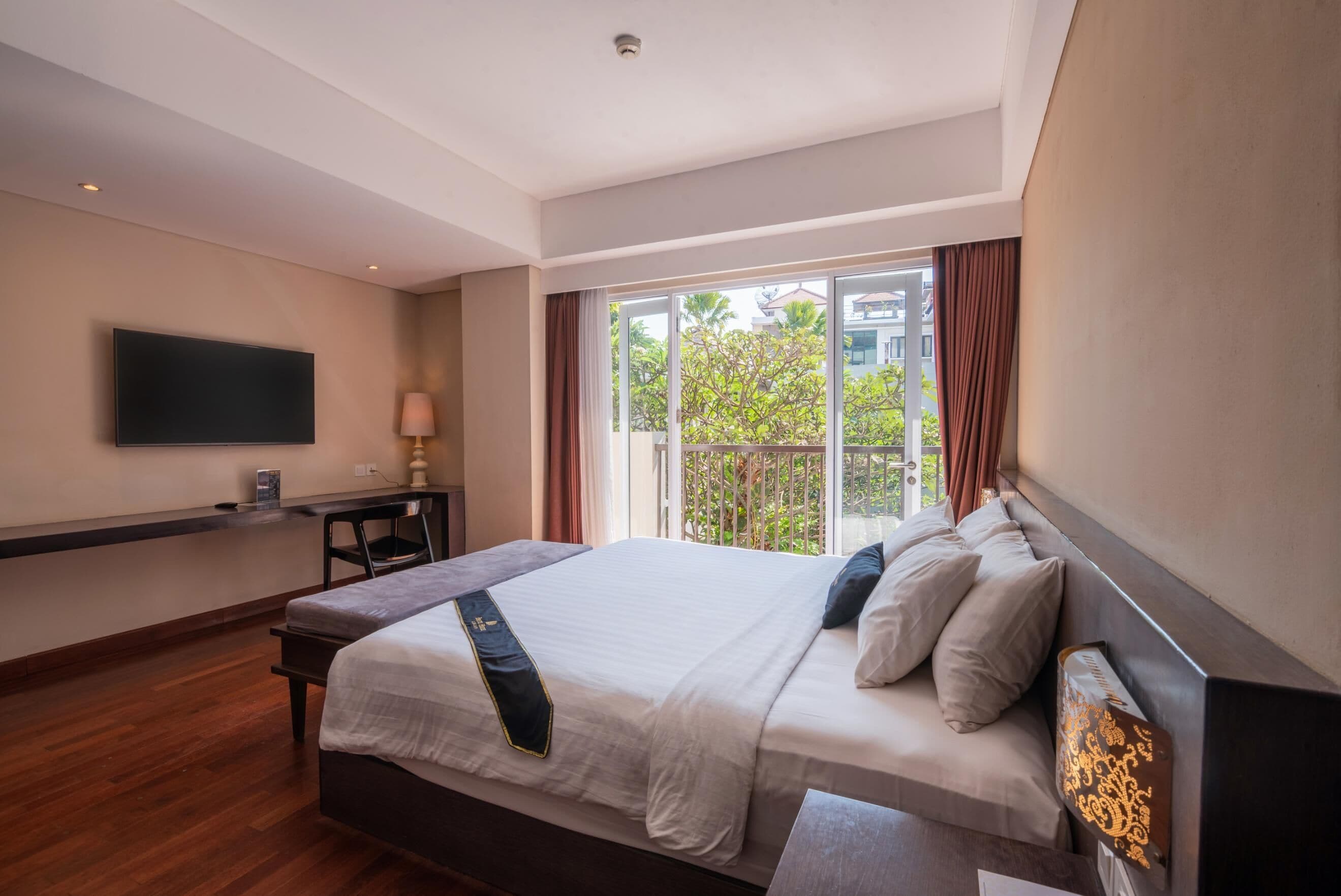 A hotel room with a large bed, a flat-screen TV mounted on the wall, a desk with a lamp, and a window with curtains.