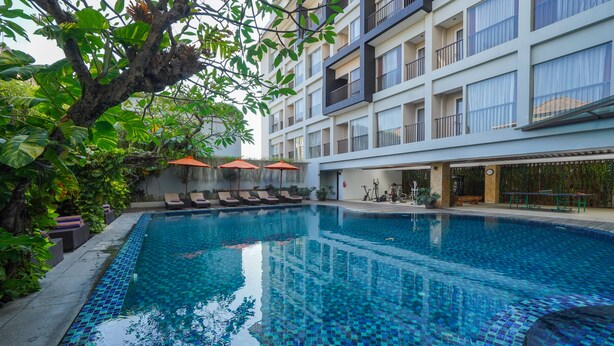 A swimming pool with a tiled edge, surrounded by lounge chairs and umbrellas, with a building in the background. - Brits Hotel Legian (Kuta)