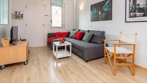 A modern living room with a grey sofa, a small white table, and a wooden chair. There is a cart with a box and a picture frame on the floor. - Duplex Cosy avec Grande Terrasse à 50m de la Plage de Palamós (Palamós)