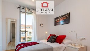 A modern bedroom with a bed, nightstands, and a view of buildings through a large window. - Duplex Cosy avec Grande Terrasse à 50m de la Plage de Palamós (Palamós)