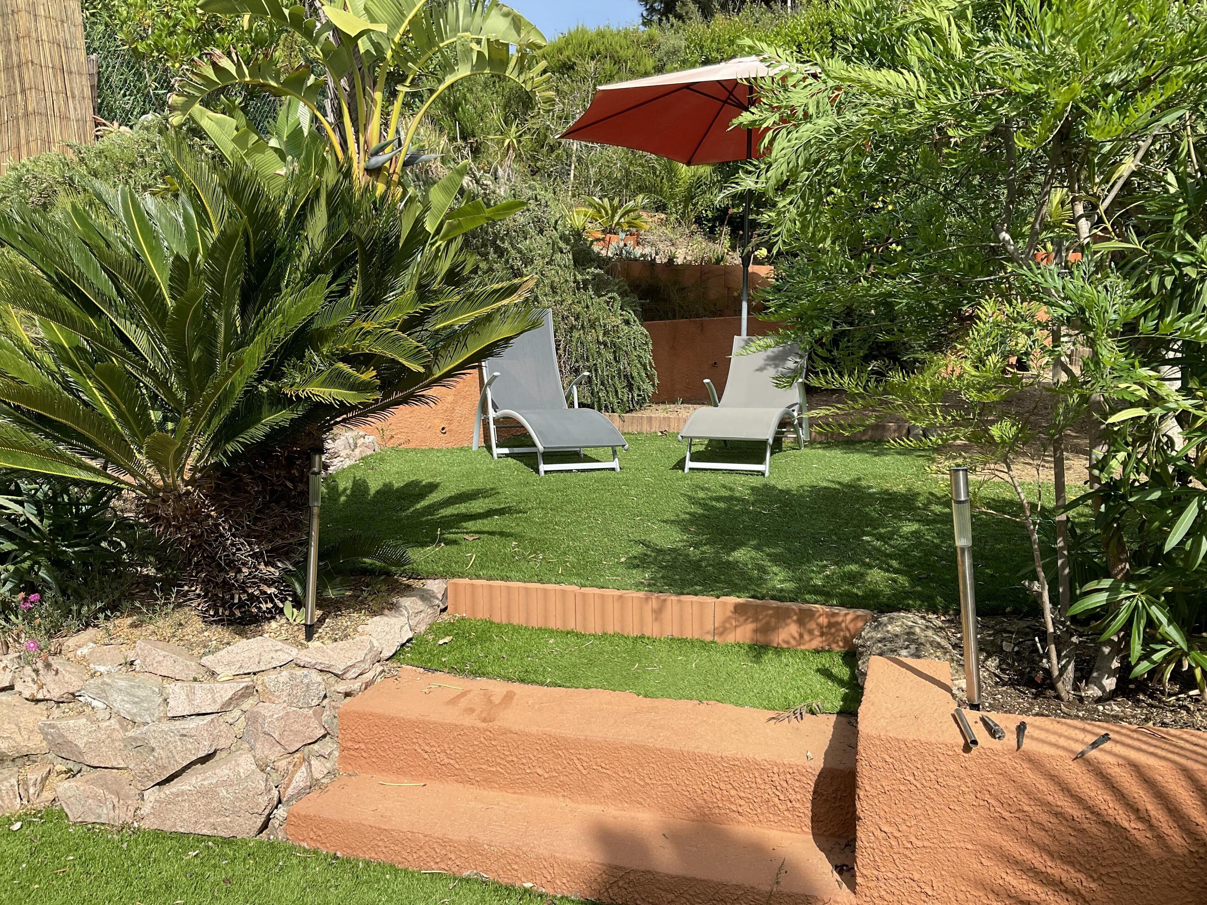 Un jardin avec deux chaises longues blanches, un parasol rouge et une variété de plantes.