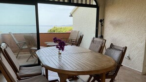 Une table en bois avec six chaises, un vase rempli de fleurs violettes et une vue sur la mer, visible à travers une porte vitrée.