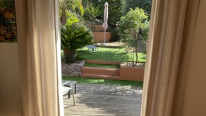 Une vue à travers une fenêtre avec des rideaux, dévoilant une terrasse en bois, un petit jardin avec une pelouse, un mur de pierre et une table en métal.