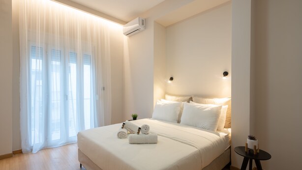 Ein modernes Hotelzimmer mit einem großen Bett, einem kleinen Beistelltisch und einem Fenster mit transparenten Vorhängen.