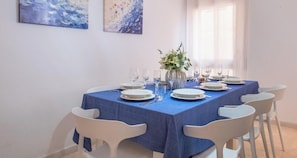 Une table à manger dressée pour six personnes, avec des chaises blanches, une nappe bleue et un vase rempli de fleurs.