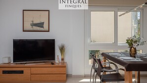 Ett modernt vardagsrum med en platt-TV placerad på ett träskåp, ett matbord med stolar och ett stort fönster som släpper in naturligt ljus.