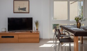 Un salon moderne avec un téléviseur à écran plat posé sur un meuble en bois, une table à manger avec des chaises et une grande fenêtre laissant entrer la lumière naturelle.