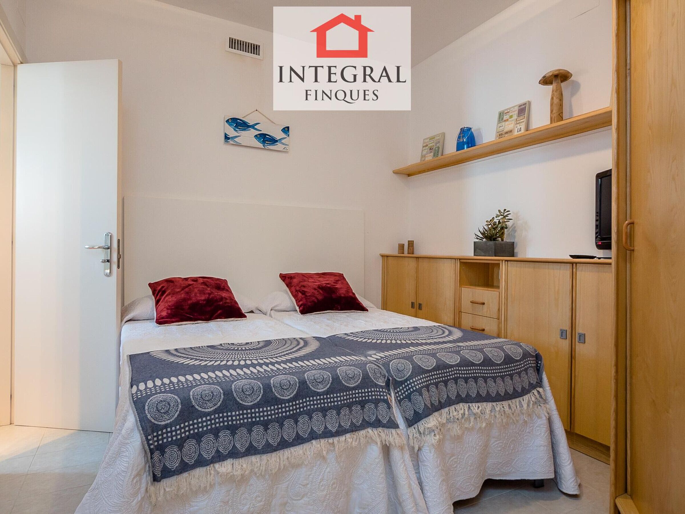 Un dormitorio con una cama, dos almohadas rojas, un edredón estampado, un armario de madera y una estantería con objetos decorativos.