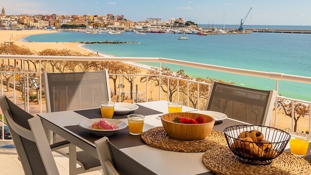 A table set with breakfast items, including fruit and juice, overlooking a coastal view with boats and buildings.