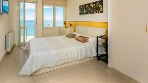 A bedroom with a bed, a nightstand, a wall art piece, and a view of the sea through a door.