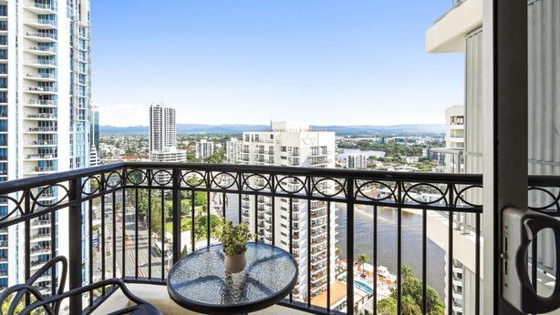 Un balcon avec une table en verre et un pot de fleurs, donnant sur un paysage urbain avec de hauts immeubles et une rivière.